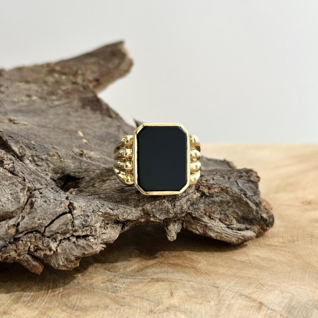 Herrenring aus 585er Gelbgold mit schwarzer Onyxplatte, Nahaufnahme auf dunklem Holzuntergrund, Fokus auf die gewölbte Ringschiene und den ruhigen Kontrast zwischen Gold und tiefem Schwarz.