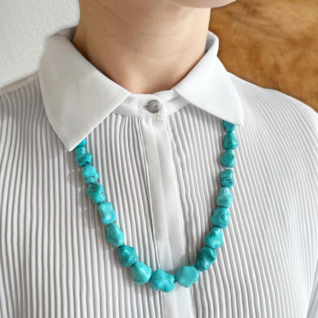 Türkis-Kette mit größeren Steinen nach vorne hin verlaufend, 60cm lang, getragen zu einer eleganten weißen Bluse, sodass Farbe und natürliche Struktur der Steine stilvoll zur Geltung kommen.