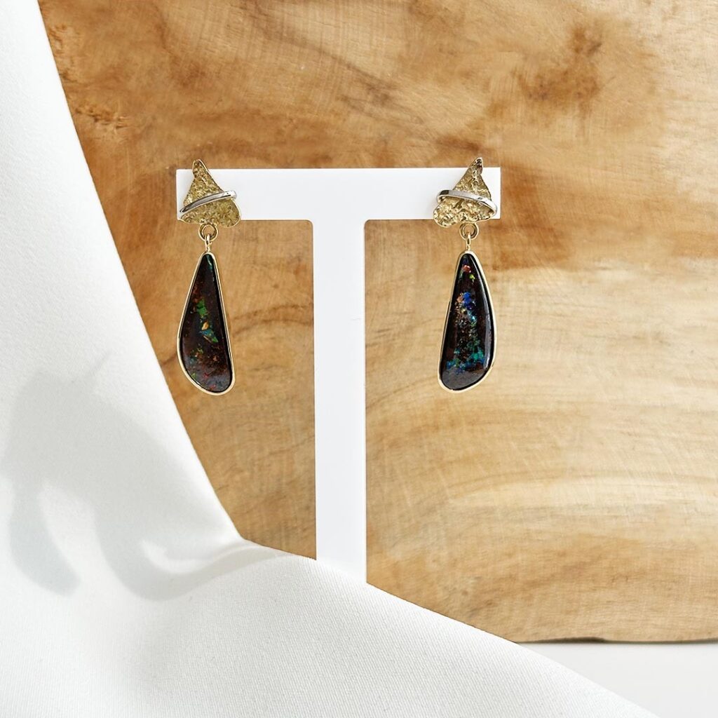 Handgefertigte Ohrringe aus 585 Gelb- und Weißgold mit Matrix Boulder Opal vor einer Holzwand präsentiert.