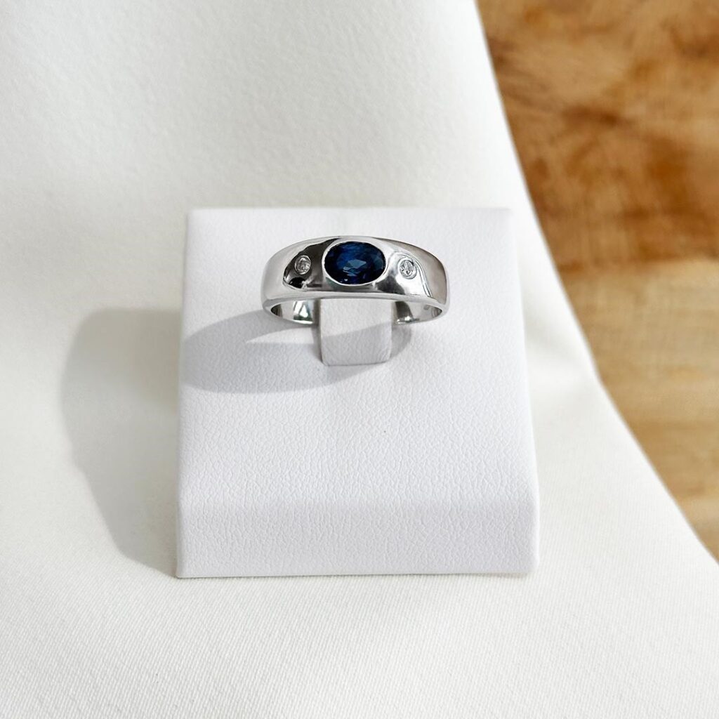 Klassischer Allianzring aus 585er Weißgold mit ovalem blauem Saphir in der Mitte und zwei seitlich eingefassten Brillanten, elegant frontal auf einem Ringpodest präsentiert; klare Linien und edle Farbkontraste wirken besonders harmonisch.