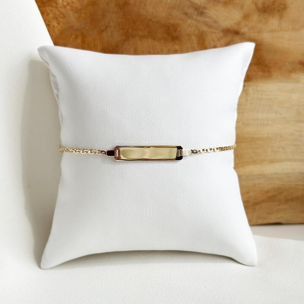 Schildarmband aus 585er Gelbgold mit zarter Broadkette, gravierbarem Schild (25×5 mm) und Zwischenöse bei 12cm, liebevoll als klassisches Tauf- und Geburtsgeschenk arrangiert.
