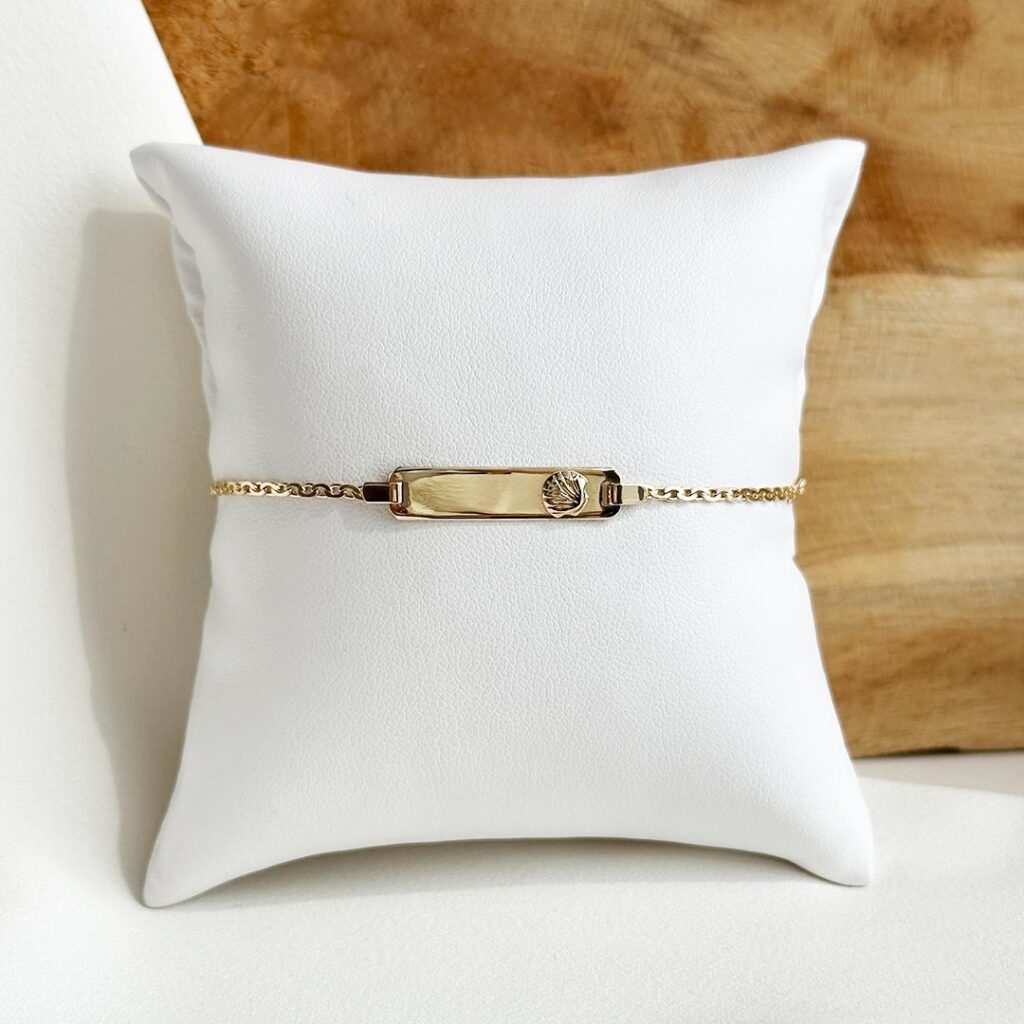 Schildarmband aus 585er Gelbgold mit feiner Brotkette, kleinem Muschel-Motiv auf dem gravierbaren Schild und Zwischenöse bei 12cm, liebevoll arrangiert für ein elegantes Produktfoto.