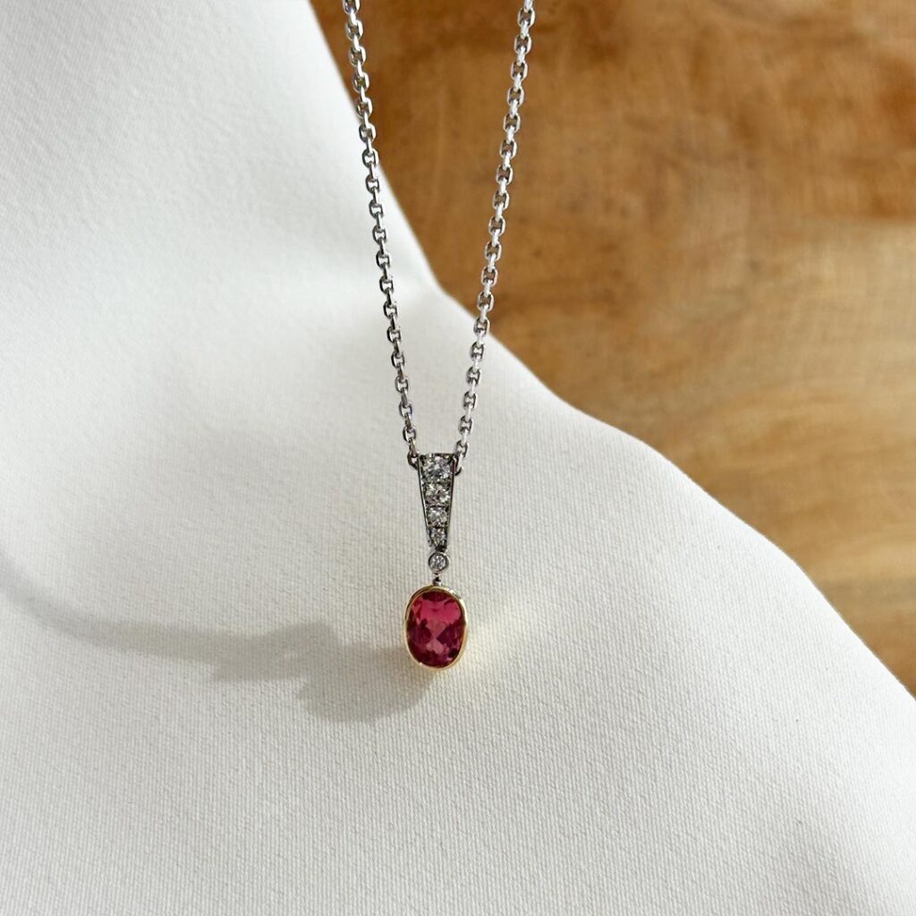 Handgefertigter Anhänger aus 585 Weiß- und Gelbgold, 20mm groß, mit abgestuft gefassten Brillanten im Weißgold-Bereich und ovalem rosa-rot schimmerndem Turmalin in glatter Gelbgoldfassung, in eleganter Nahaufnahme auf einer Weißgoldkette präsentiert.