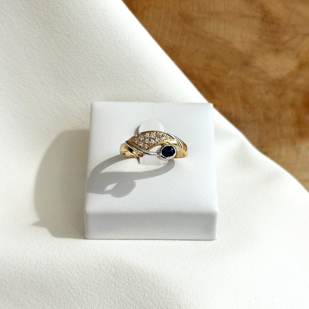 Zweifarbiger Ring aus 585er Gelb- und Weißgold mit Brillantreihe und zentralem ovalen Saphir, in klarer Frontansicht auf einem Ringpodest elegant fotografiert.