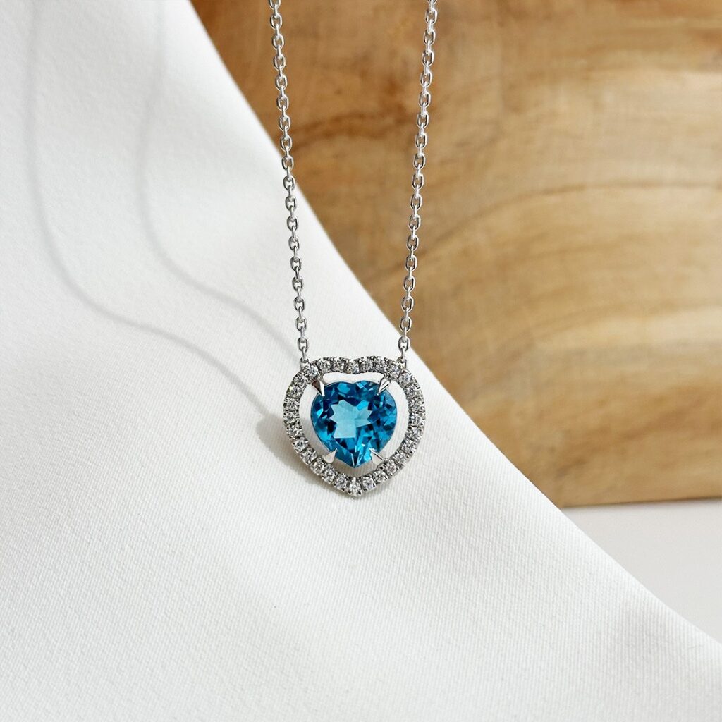 Collier aus 585 Weißgold mit herzförmigem blauem Topas und diamantgefasstem Herzrahmen fotografiert vor einer Holzwand.