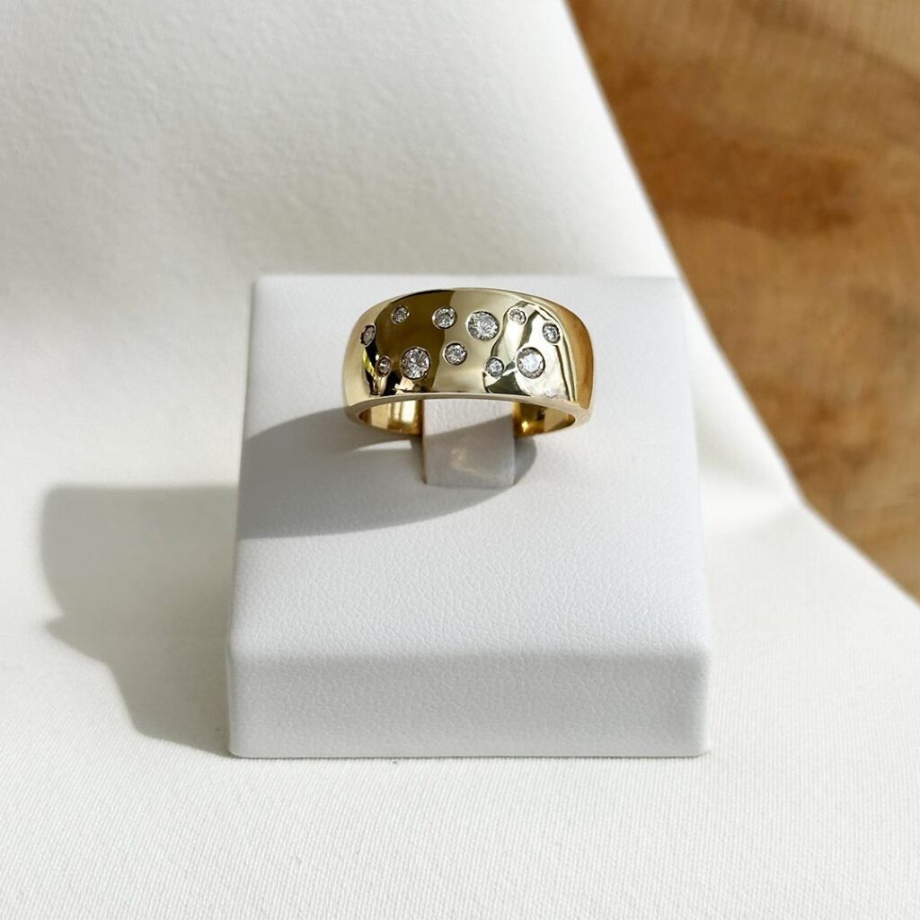 Breiter 585-Gelbgoldring mit polierter Oberfläche auf einem Ringpodest; elf unterschiedlich große Brillanten wirken wie verstreut über die Ringoberseite und erzeugen einen lebendigen, funkelnden Effekt.