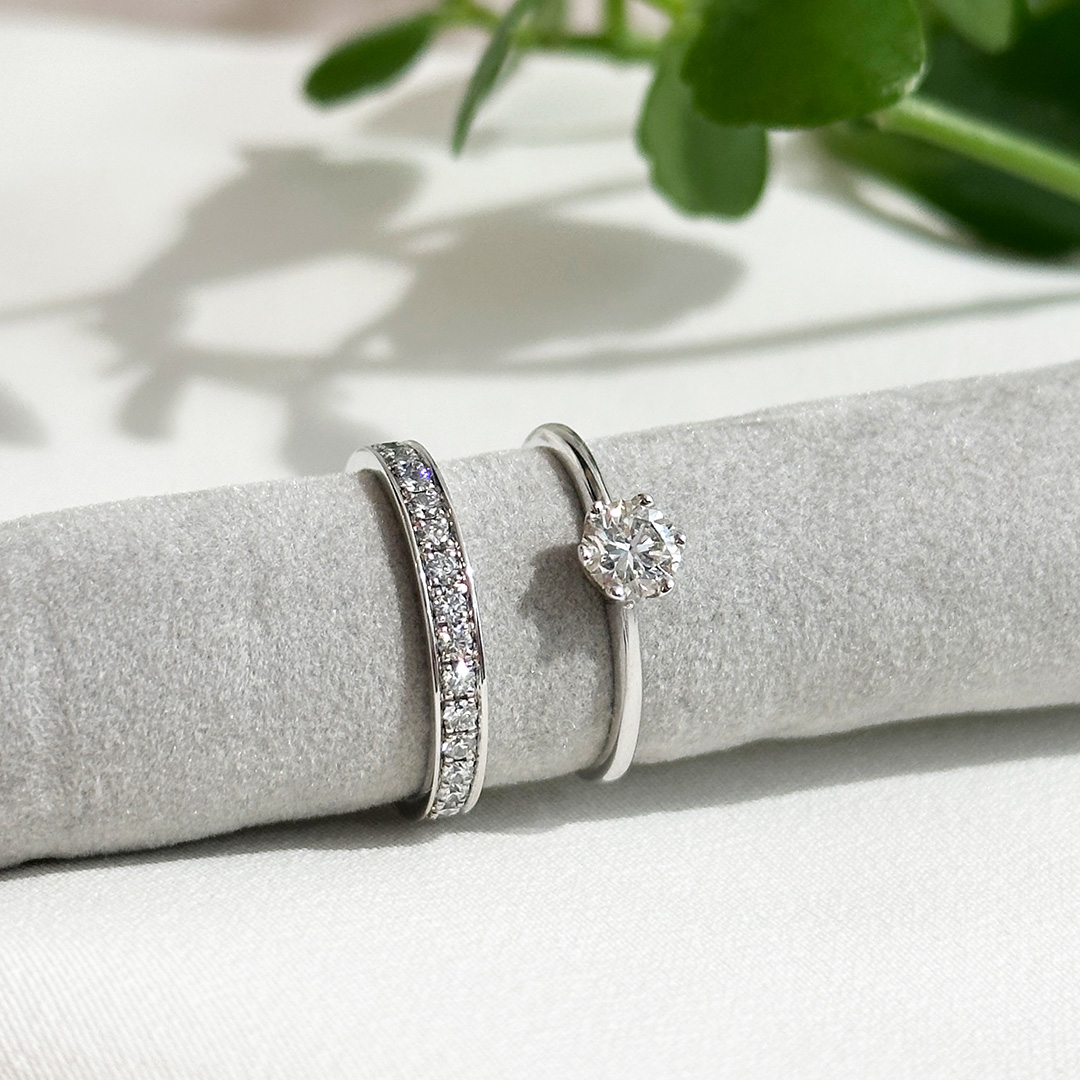 Solitärring aus Weißgold mit klassischer Sechskrappenfassung kombiniert mit Diamantring, auf grauem Präsentationsstoff mit dezenten Pflanzenelementen im Hintergrund, Fokus auf die elegante Ringkombination und die feine Brillanz