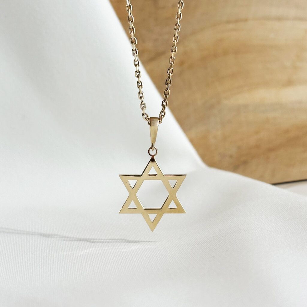 Davidstern-Anhänger aus 585er Gelbgold, 17mm groß, hängend an einer feinen Goldkette vor einer eleganten Holzwand fotografiert, Fokus auf die offene Gestaltung des Sterns und seine symbolische Bedeutung für Glauben, Identität und Schutz, zeitloser Schmuck mit besonderer Aussagekraft.