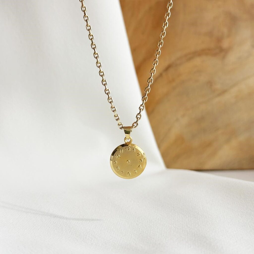 Die Taufuhr aus 585er Gelbgold, 12mm groß, hängend an einer feinen Goldkette vor einer Holzwand fotografiert, Fokus auf den polierten Rand und das individuelle Uhrmotiv mit gravierter Geburtszeit, persönliches Geschenk zur Geburt oder Taufe mit Platz für eine Gravur auf der Rückseite.
