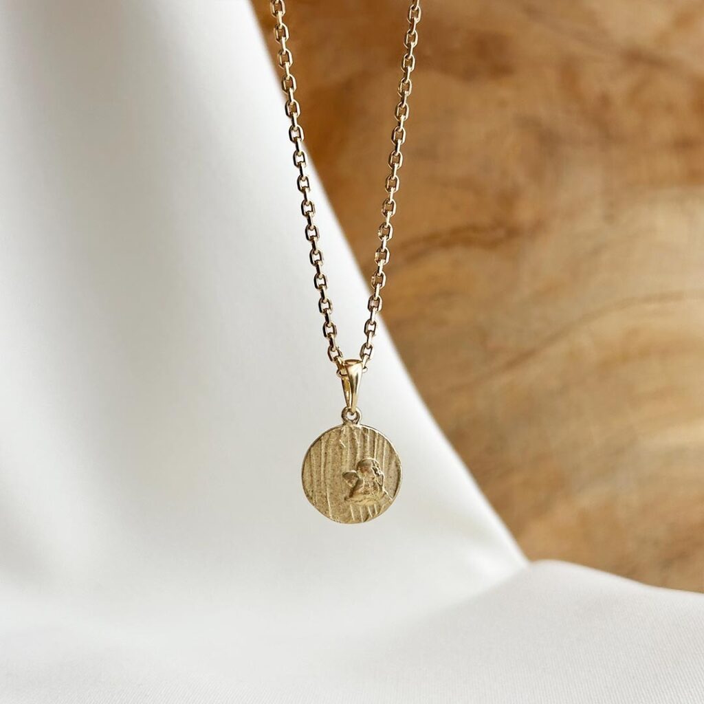 Der Schutzengel-Anhänger aus 585er Gelbgold, 11mm groß, hängend an einer feinen Goldkette vor einer Holzwand fotografiert, Fokus auf die markant strukturierte Oberfläche, das klassische Engelmotiv und die Möglichkeit einer persönlichen Gravur auf der Rückseite, bedeutungsvolles Geschenk zur Geburt, Taufe oder Erstkommunion