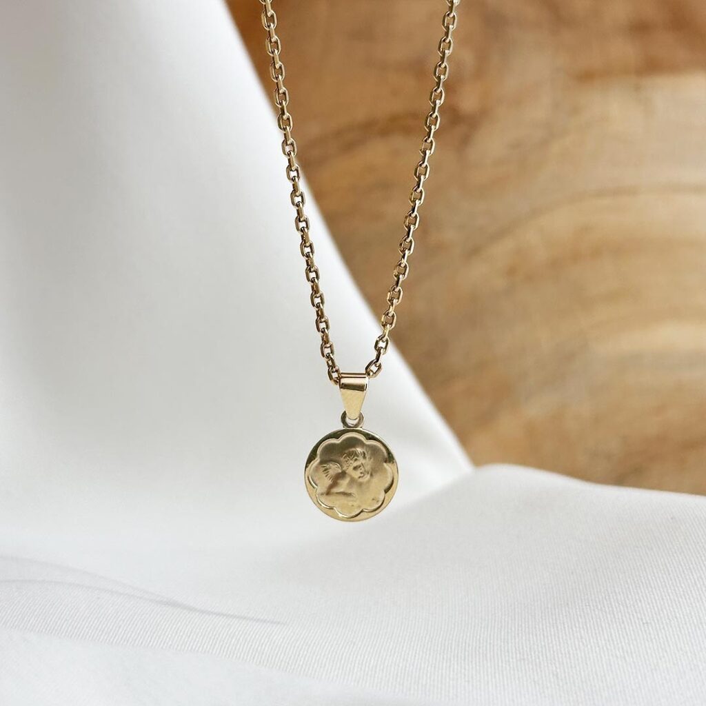 Der Schutzengel-Anhänger aus 585er Gelbgold, 10mm groß, hängend an einer feinen Goldkette vor einer Holzwand fotografiert, Fokus auf die blütenförmige Umrandung, den liebevoll gestalteten Engel und die symbolische Bedeutung als Geschenk zur Geburt, Taufe oder Erstkommunion