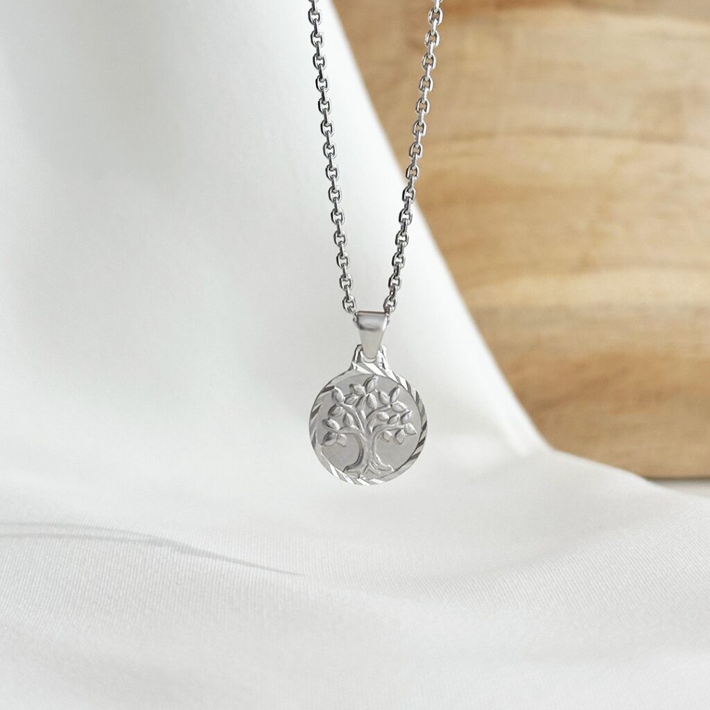 Lebensbaum-Anhänger mit facettierter Umrandung, hängend an einer feinen Goldkette vor einer eleganten Holzwand fotografiert, Fokus auf den detailreich gearbeiteten Lebensbaum, den mattierten Hintergrund und die lichtreflektierende Umrandung, symbolträchtiger Schmuck für Wachstum, Stärke und Verbundenheit