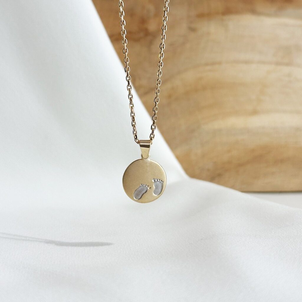 Der Anhänger mit Babyfüßen aus 585er Gelb- und Weißgold, hängend an einer feinen Goldkette vor einer Holzwand fotografiert, Fokus auf die zweifarbige Gravurplatte mit den kleinen Babyfüßen und die persönliche Symbolik als Geschenk zur Geburt, zum Muttertag oder als bleibende Erinnerung an einen besonderen Lebensmoment