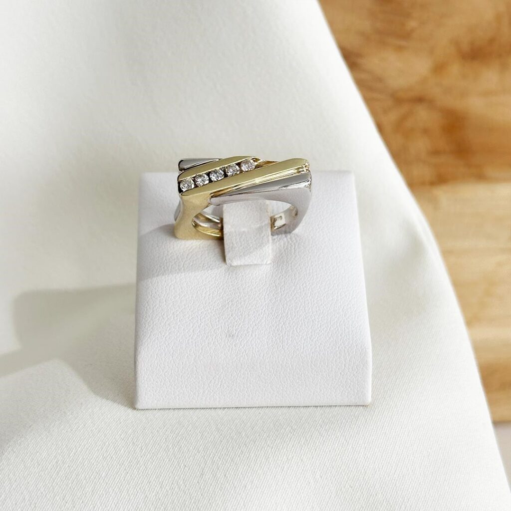 Bicolor-Ring aus 585er Gelb- und Weißgold mit fünf Brillanten, frontal auf einem Ringpodest fotografiert, Fokus auf die markante eckige Form, das Zusammenspiel der beiden Goldfarben und die harmonisch gefassten Brillanten, die dem Ring ein feines Funkeln verleihen