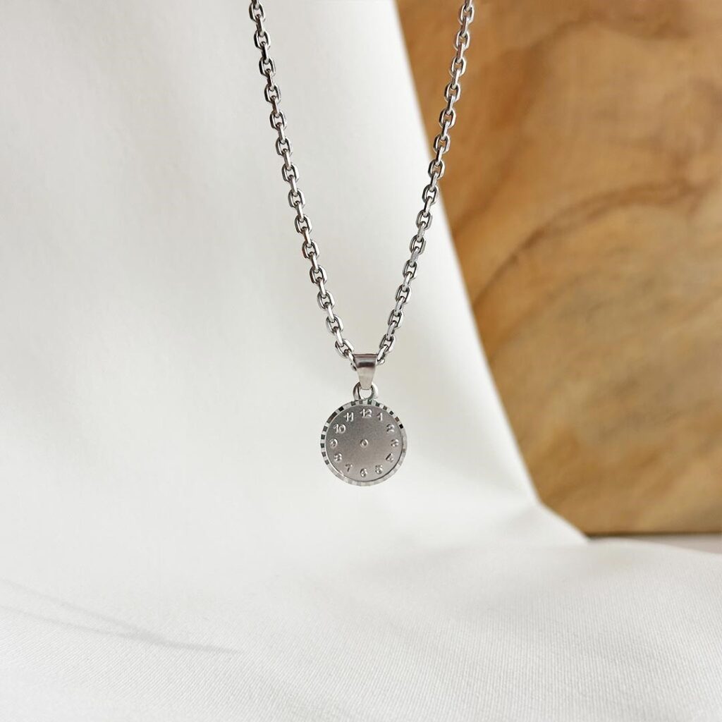 Die Taufuhr aus 585er Weißgold, 10mm groß, hängend an einer feinen Weißgoldkette vor einer Holzwand fotografiert, Fokus auf das individuelle Uhrmotiv mit gravierter Geburtszeit und die zeitlose Gestaltung, persönliches Geschenk zur Geburt oder Taufe mit Möglichkeit zur Gravur auf der Rückseite